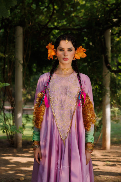 A woman stands outdoors in a wooded setting wearing an ornate lavender traditional dress with gold embroidery, bright orange flower headpieces, and green sleeves. The background consists of blurred trees and light posts.