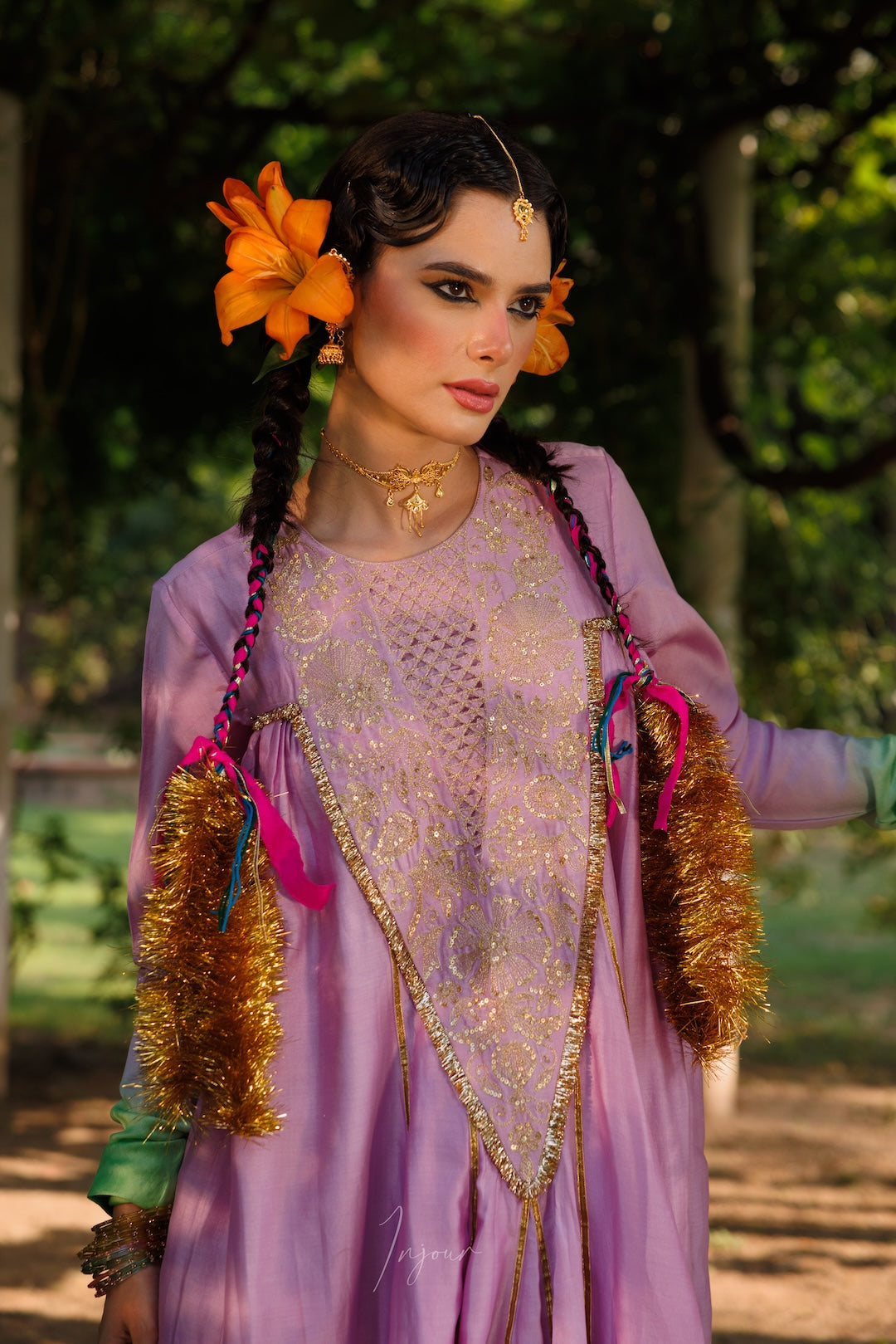 A woman wearing a traditional lavender and gold embroidered dress with orange flowers in her hair, standing outdoors in a park-like setting with trees in the background.