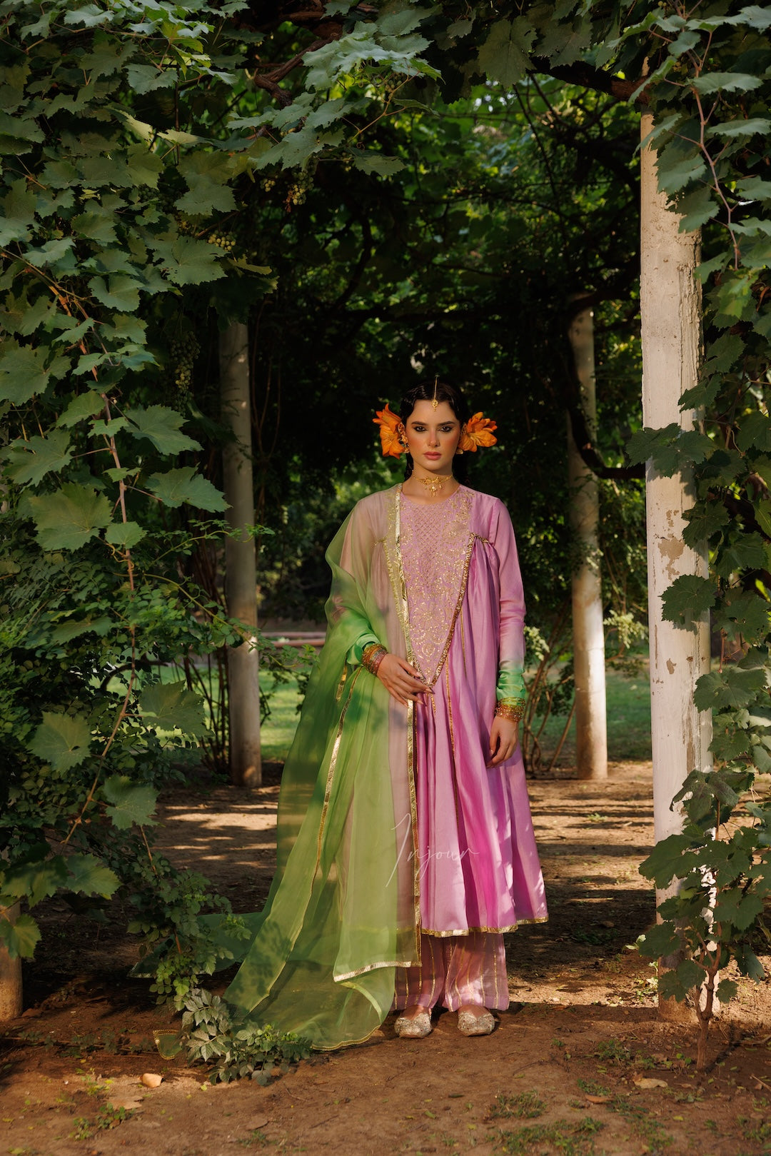 A woman stands outdoors in a garden wearing a traditional outfit consisting of a pink embroidered kurta, matching pants, and a sheer green dupatta draped over one side.
