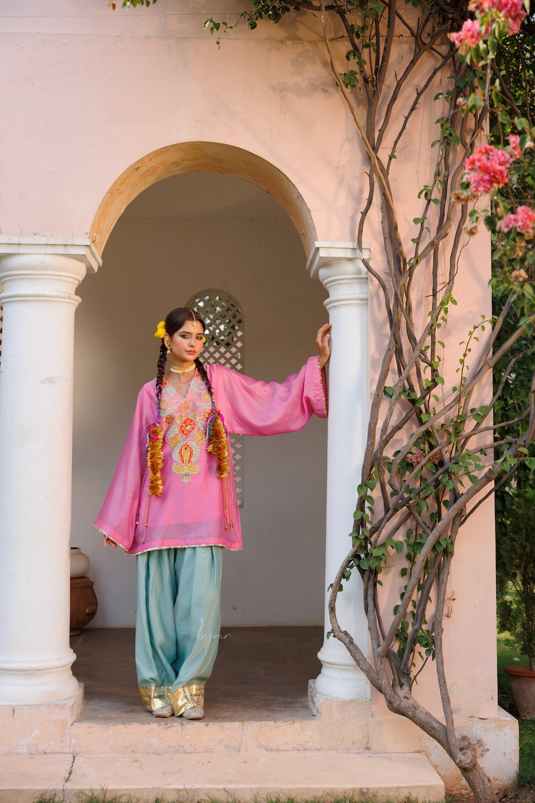 A woman wearing a traditional pink embroidered kurta with light blue pants and gold shoes, standing in a doorway of a pink building with climbing flowers.