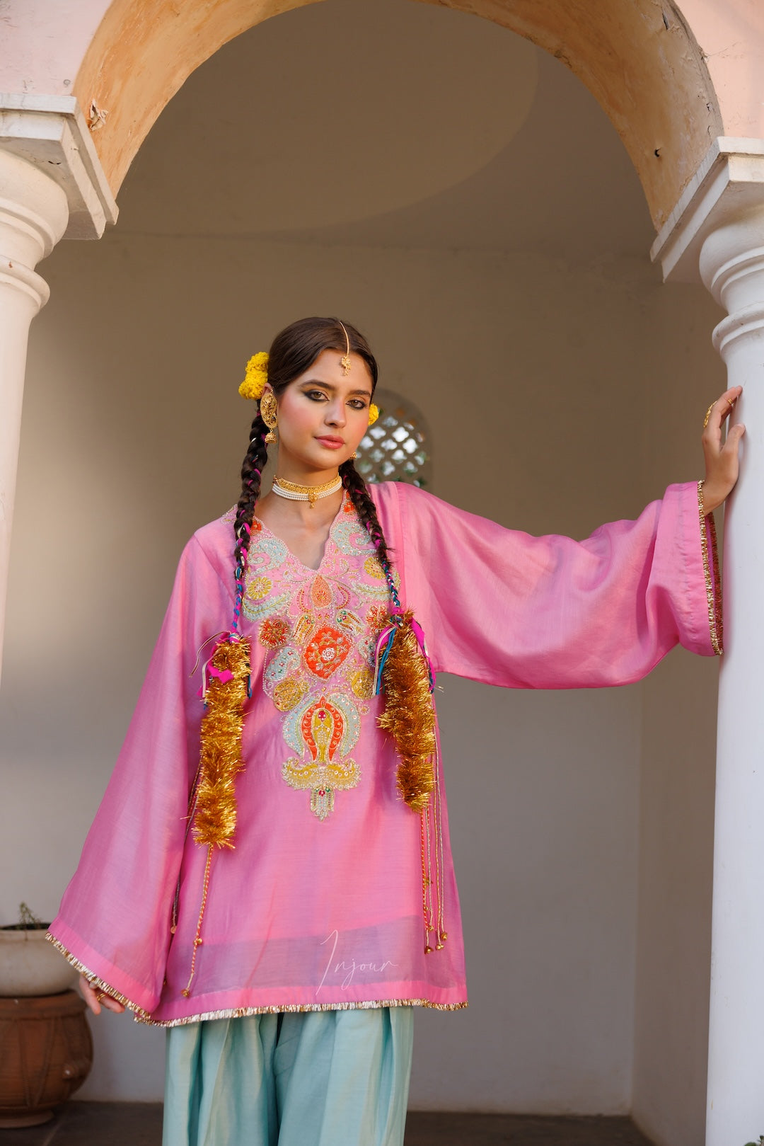 A woman models a traditional pink salwar kameez suit with intricate gold, orange, and green embroidery on the chest, paired with light blue pants. She is standing in a courtyard framed by white pillars and an arched doorway.