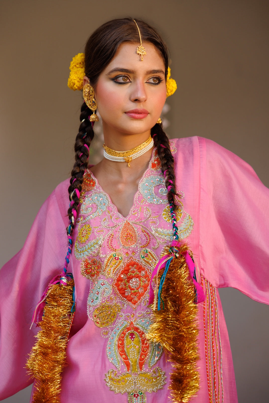 A woman wearing a bright pink embroidered traditional outfit with colorful floral patterns, gold jewelry, braided hair with pink and blue threads, and yellow hair accessories, set against a plain warm brown background.