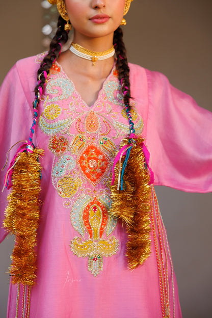A close-up of a model wearing a vibrant pink traditional embroidered dress with intricate multicolor beadwork and gold tinsel fringe on the chest, styled with gold jewelry.