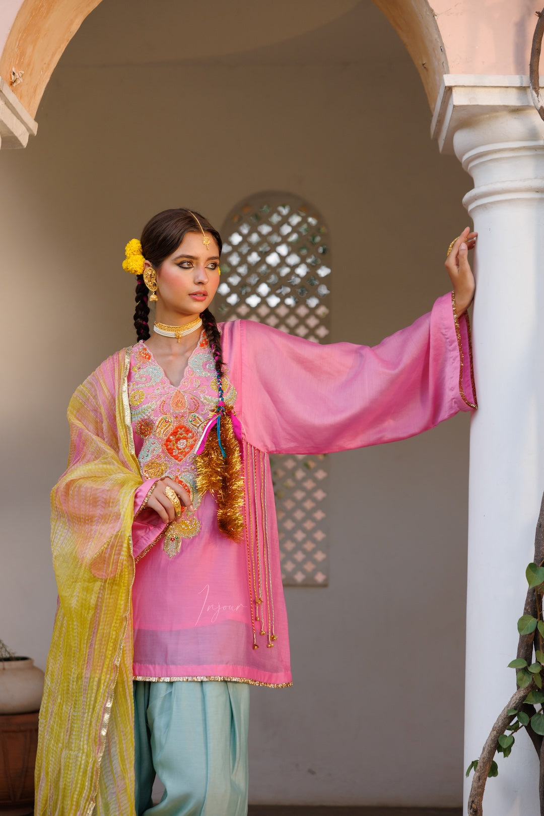 A woman modeling a traditional pink salwar kameez outfit with a yellow dupatta, light blue pants, and gold jewelry, standing in an arched corridor.