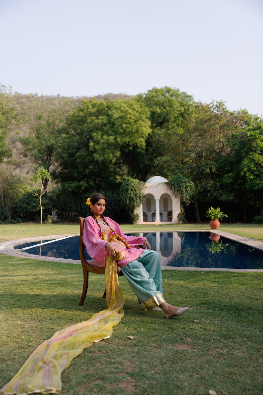 A woman wearing a vibrant pink and teal traditional outfit with a yellow dupatta sits on a wooden chair in a lush garden setting with a swimming pool and a small white pavilion in the background.