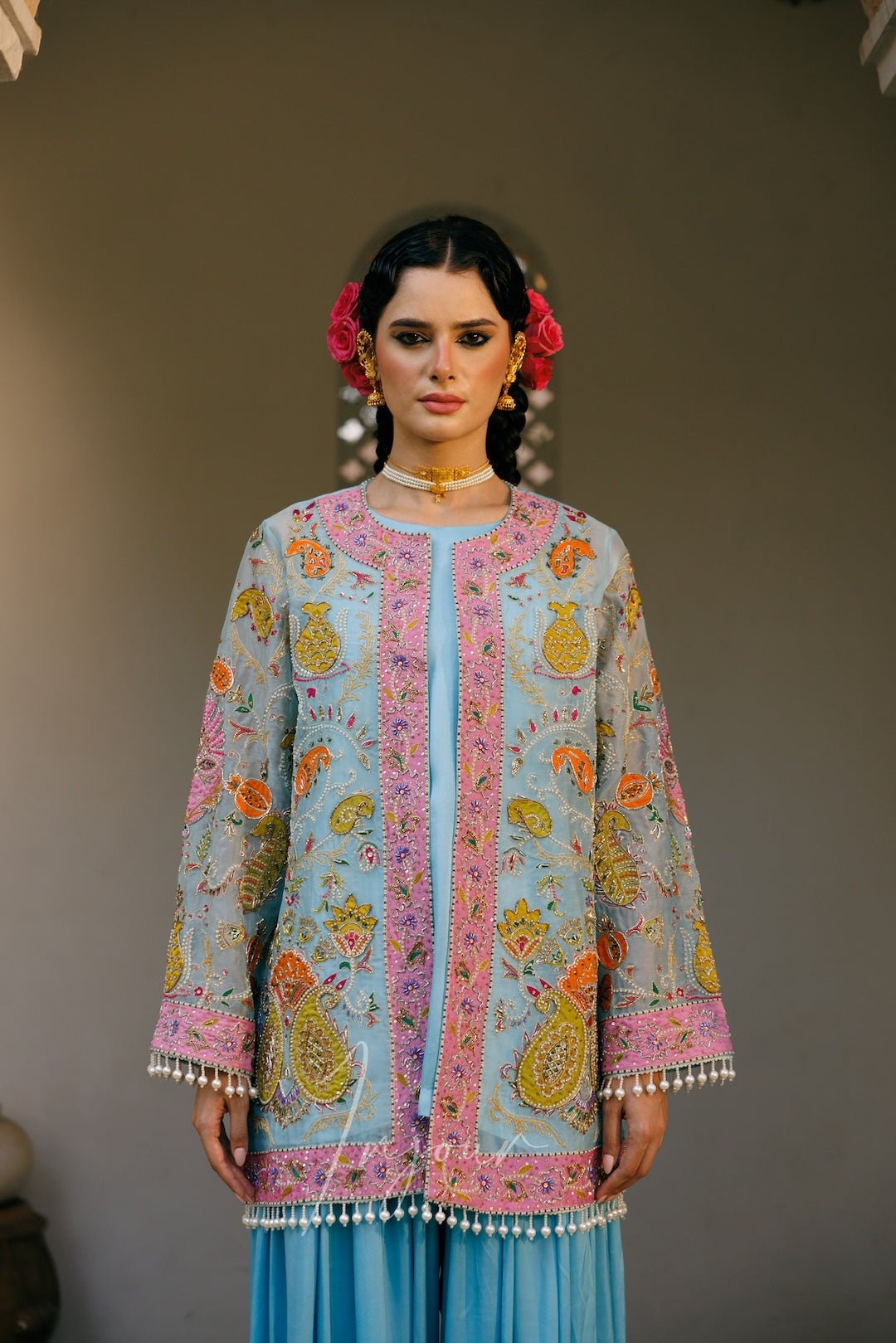 A woman modeling a light blue embroidered traditional kurta with pink borders and multicolor floral motifs, paired with a matching light blue skirt, set against a neutral gray background.
