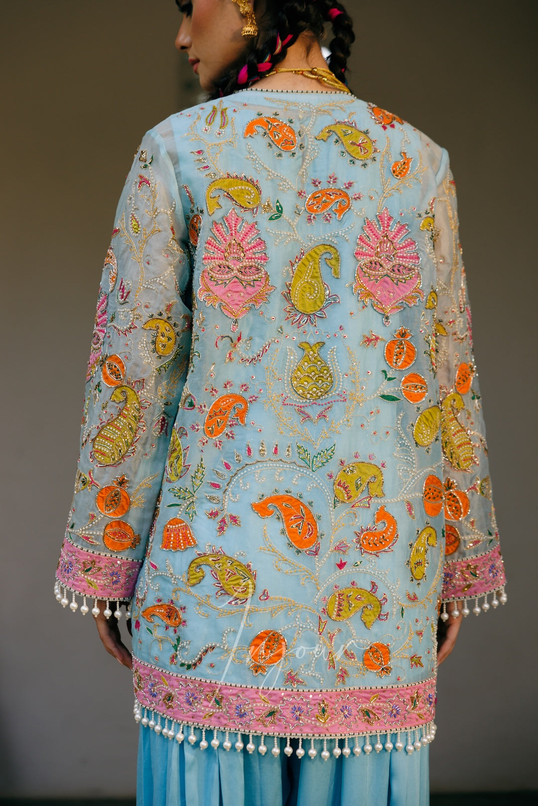 A woman shown from the back wearing a light blue embroidered kurta with colorful paisley and floral motifs in pink, orange, yellow, and green, finished with a pink border and white tassel trim.