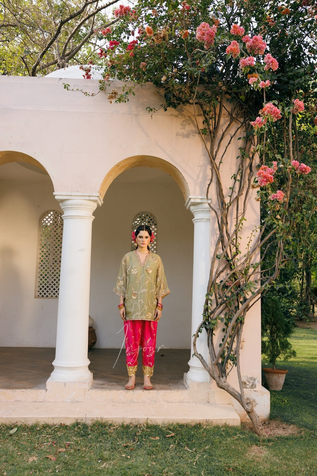A person stands in the center of a white arched pavilion with pink flowers climbing up the structure. The person is wearing a light olive-green embroidered kurta, bright pink pants, and gold boots, holding a sword.
