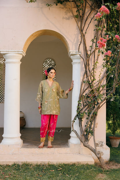 A woman stands in the center of a white arched doorway wearing a traditional outfit with a light olive green embroidered kurta and bright pink pants. Pink flowering vines climb along the right side of the wall.