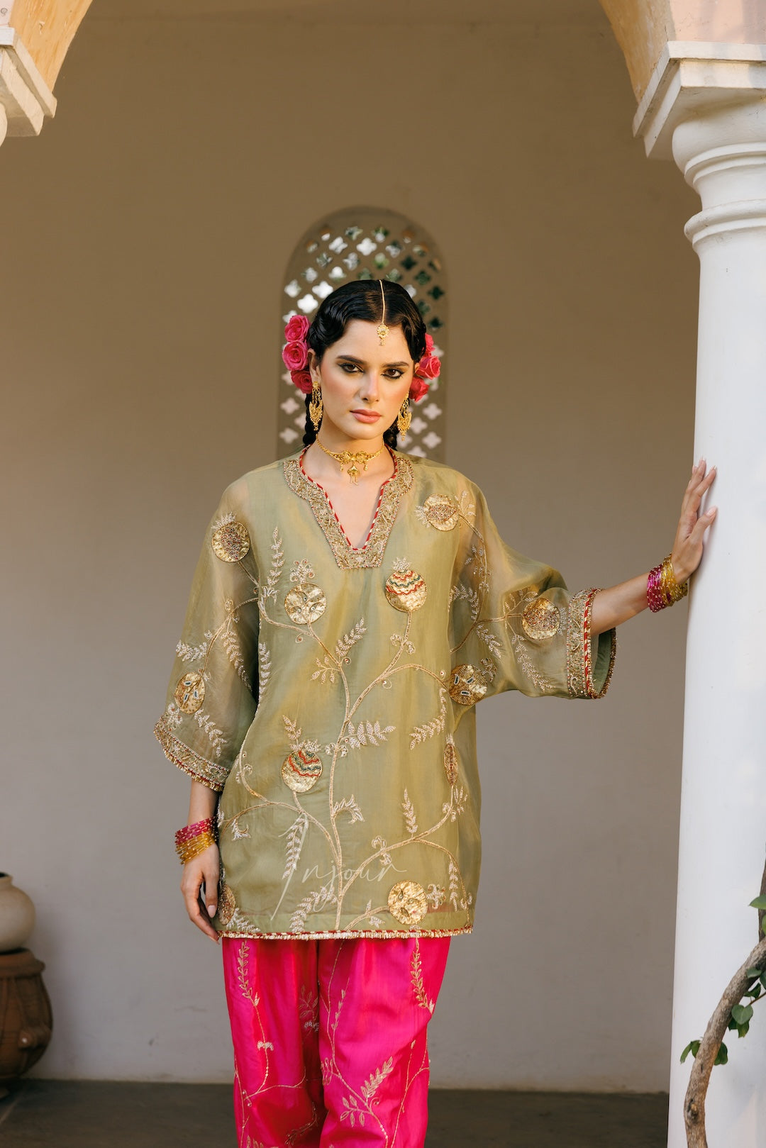 A woman modeling a traditional Indian outfit consisting of an olive green embroidered kurta and bright pink pants, standing in a doorway with white pillars.