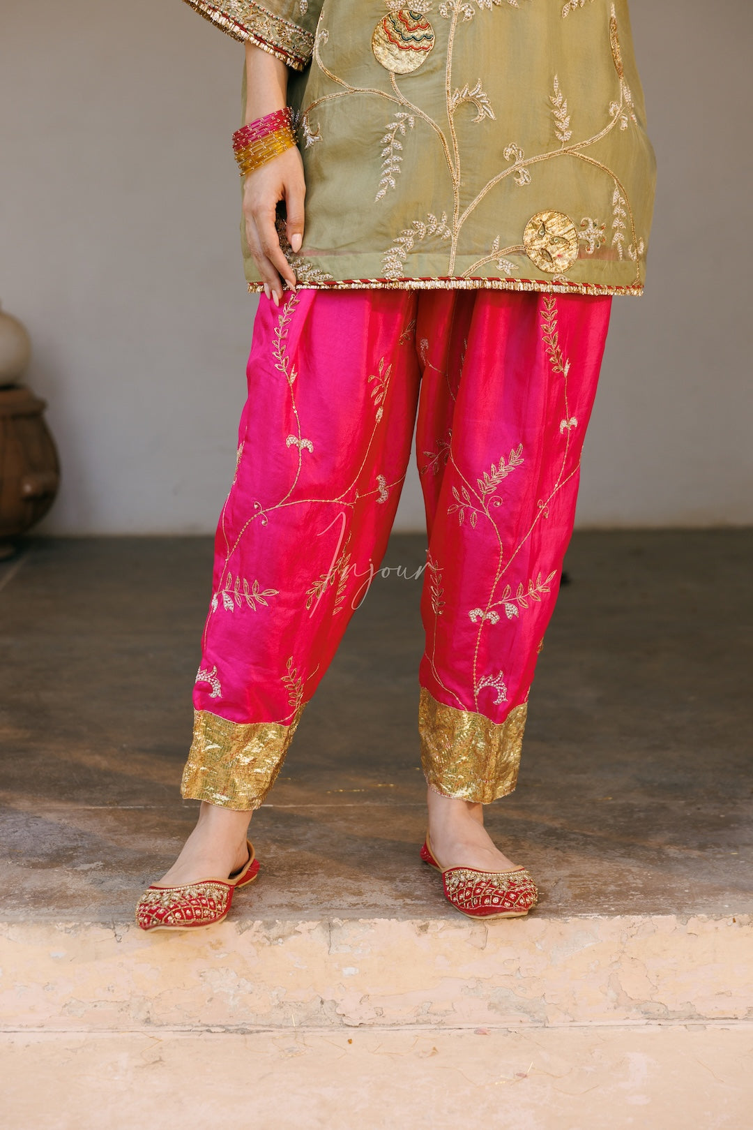 A person wearing traditional Indian attire featuring a sage green embroidered kurta and bright pink patola pants with gold borders, paired with red embroidered flat shoes.
