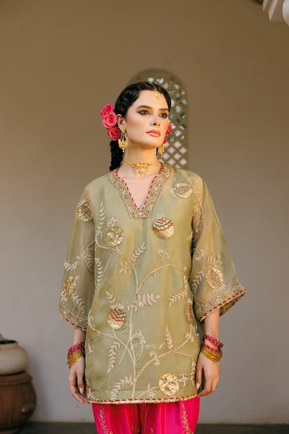 A woman models a traditional olive green embroidered kurta with gold floral patterns and bright pink pants, accessorized with gold jewelry and a pink flower in her hair, set against a neutral indoor background.