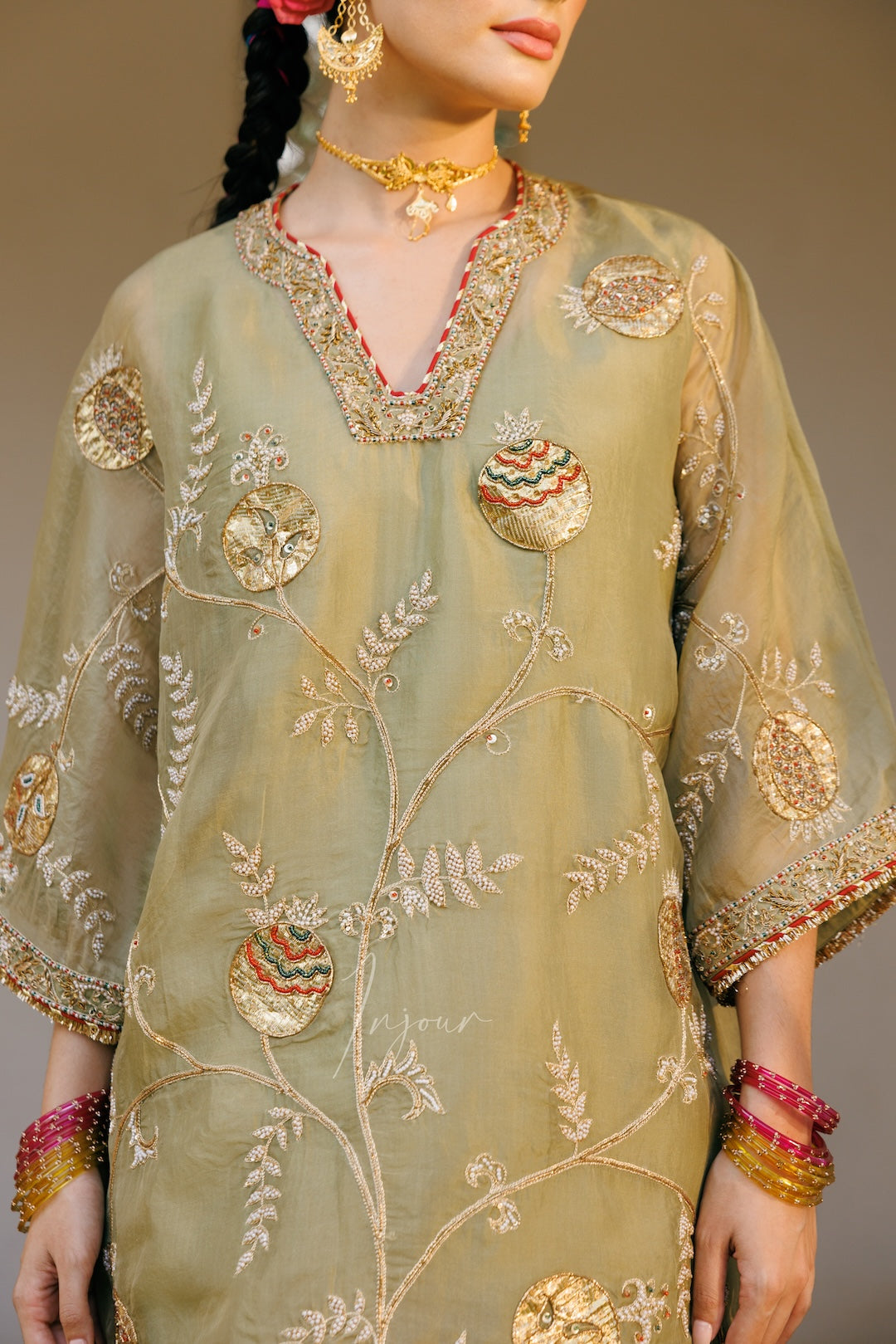 A close-up of a model wearing a sage green embroidered kurta with intricate gold, beige, and red floral patterns, styled with traditional jewelry.