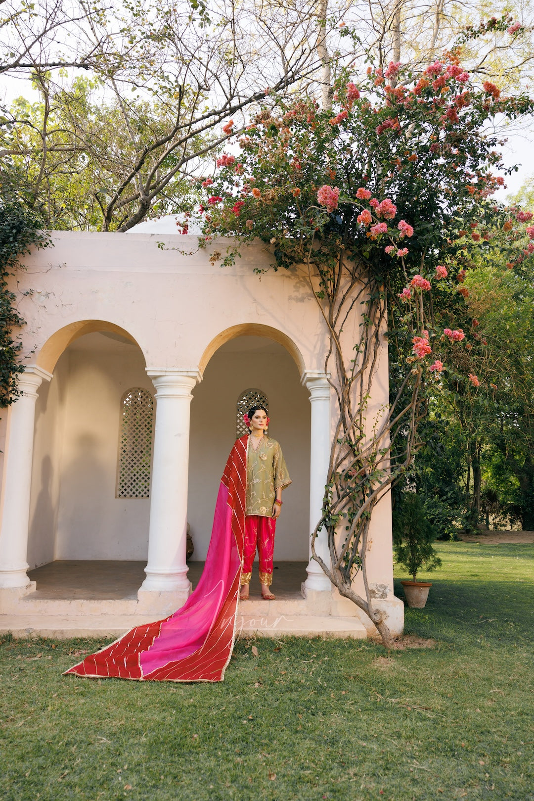 A man wearing a traditional Indian outfit stands in front of a white arched pavilion with pink flowers climbing up the side. He is wearing a beige kurta, red pants, and a bright pink and red patterned dupatta that drapes over the grass.