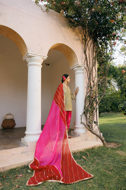 A woman stands in profile outdoors in front of a white arched pavilion, wearing a bright pink and red draped garment with gold striped borders and a beige top. Grass, trees, and garden foliage are visible around the structure.