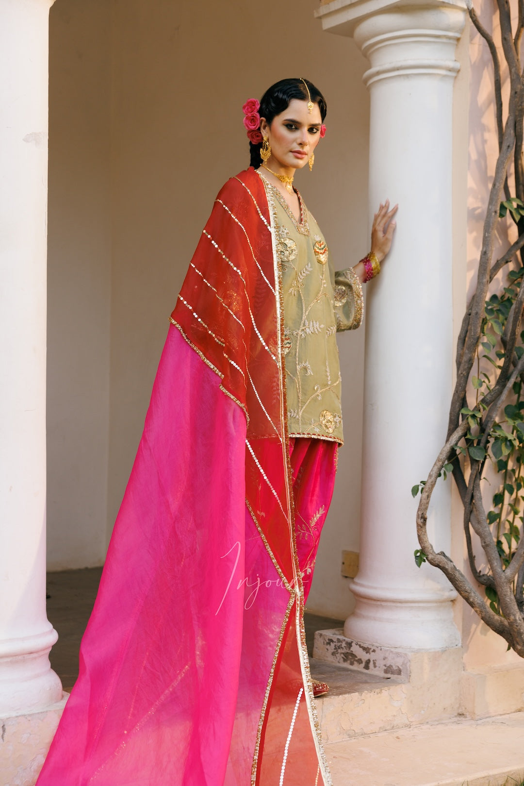 A woman models an elegant traditional outfit featuring a gold embroidered kurta, a bright pink skirt, and a red dupatta with gold borders, set against a classical white columned background.