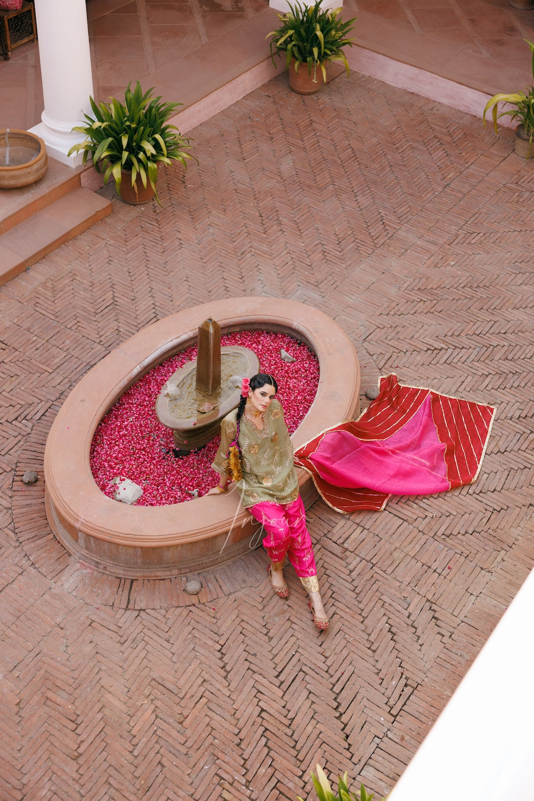 A woman in traditional attire sits beside an oval fountain in a courtyard, with a bright pink and red draped textile beside her. The setting features a brick-patterned floor and potted plants.