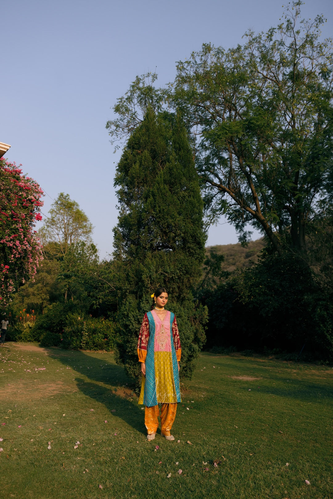 A person stands outdoors on a grassy lawn wearing a colorful traditional outfit with a pink, blue, yellow, and orange patterned tunic and bright yellow pants, surrounded by trees and shrubs under a clear sky.