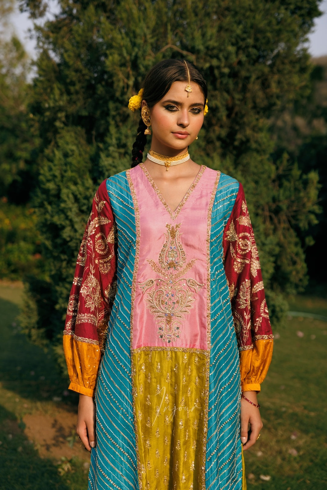 A woman wearing a traditional multicolor embroidered kurta with a pink center panel, turquoise side panels, and gold-patterned sleeves, standing outdoors in front of green trees.
