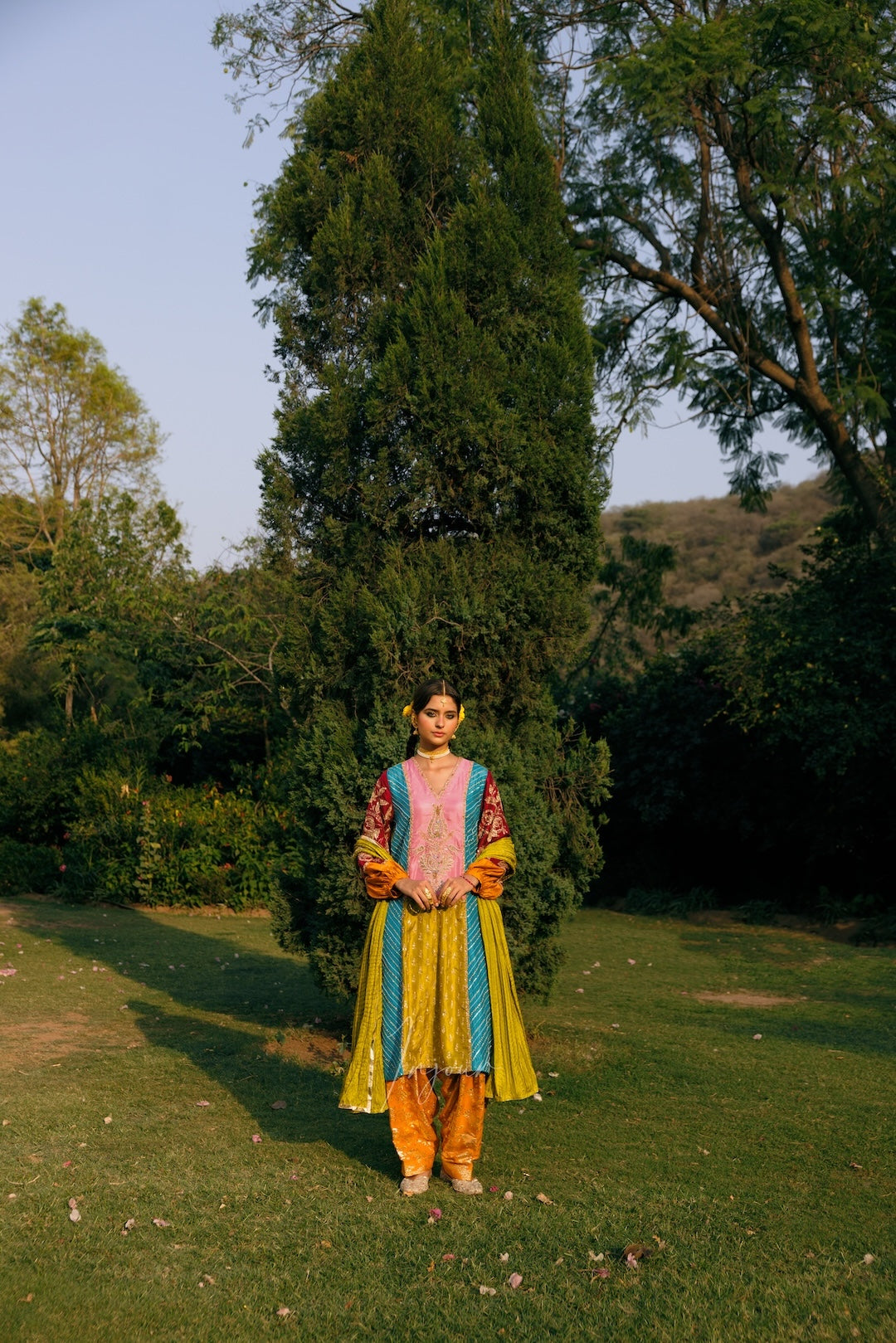 A woman stands outdoors on a grassy lawn in front of a tall evergreen tree, wearing a colorful traditional outfit with a pink top, yellow and blue skirt panels, and orange pants.