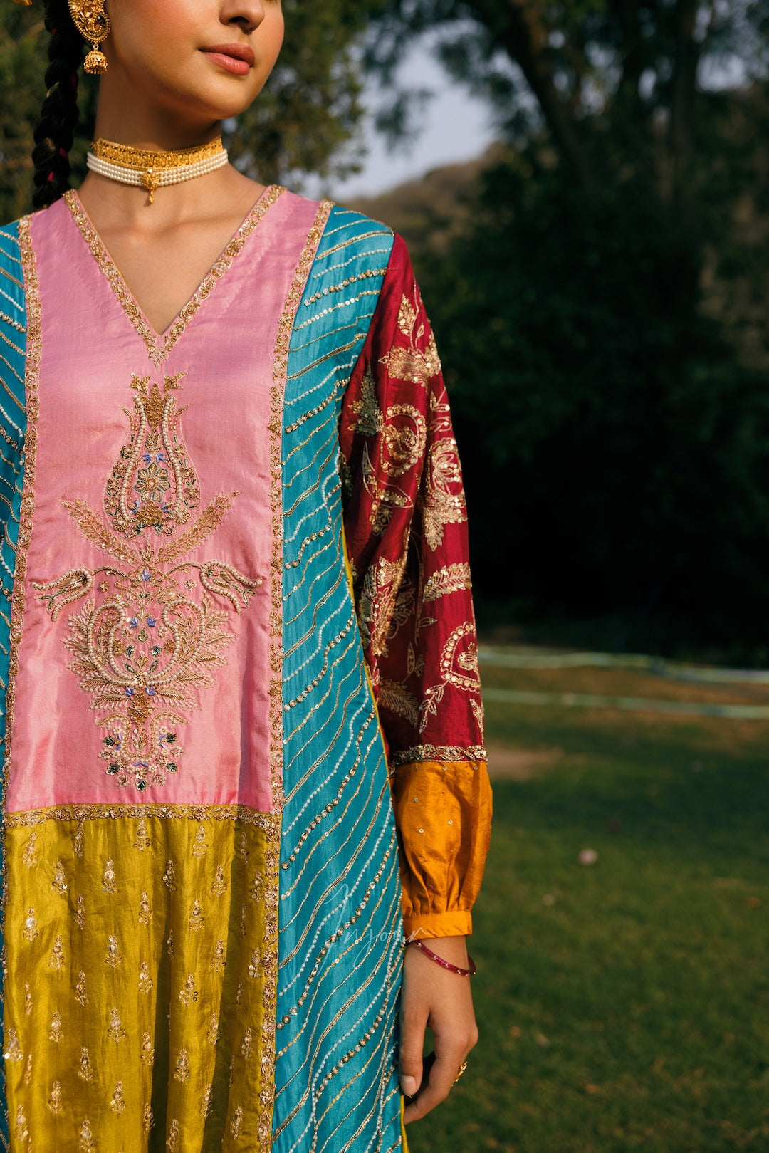 A woman outdoors wearing a traditional embroidered outfit with a pink embroidered bodice, a turquoise dupatta, and a red patterned sleeve with yellow cuffs, set against a blurred green park background.