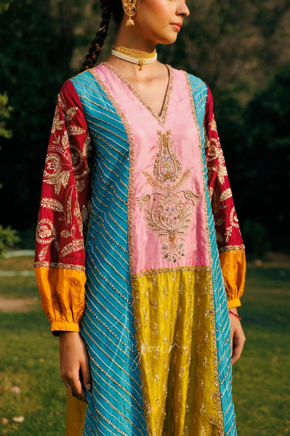 A woman wearing a colorful traditional embroidered dress outdoors. The dress has a pink V-neck bodice with gold embroidery, a yellow lower section, and red sleeves with gold patterns and yellow cuffs, accented by a turquoise striped border.