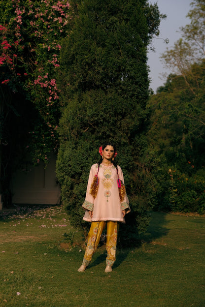 A woman stands outdoors in a garden wearing a light pink embroidered traditional outfit with yellow patterned pants. She is positioned in front of a tall, dark green tree with pink flowers on the left.