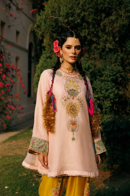 A woman stands outdoors wearing a light pink embroidered traditional outfit with yellow pants, floral hair accessories, and tassel details. The background features green trees and a building with red flowers.