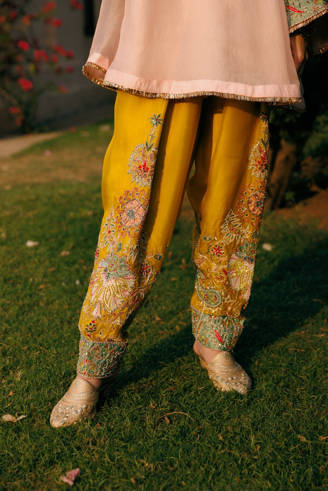 A person outdoors on grass wearing mustard yellow embroidered pants with floral and paisley patterns, a light pink top, and beige embroidered shoes.