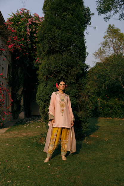 A woman stands outdoors in a garden wearing a light pink traditional outfit with gold embroidery, paired with yellow patterned pants and beige shoes. Trees, grass, and pink flowering vines are visible in the background.