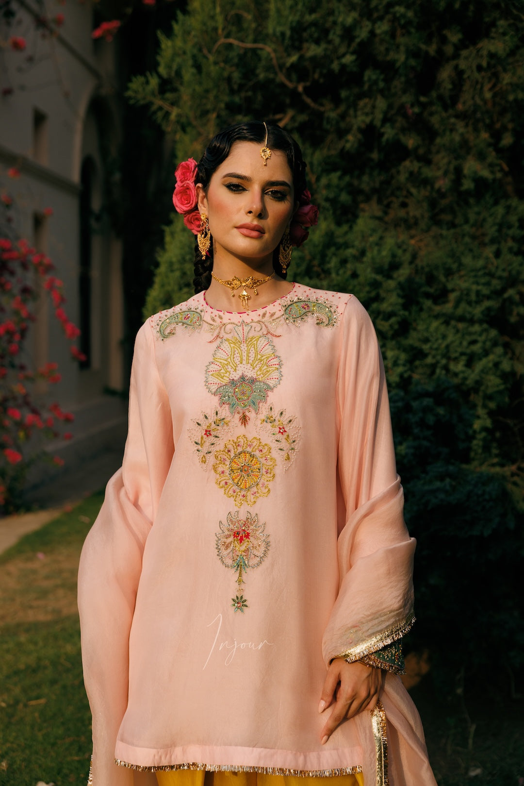 A woman wearing a light pink embroidered traditional outfit with floral patterns on the chest and sleeves, standing outdoors in front of green foliage and a stone building.