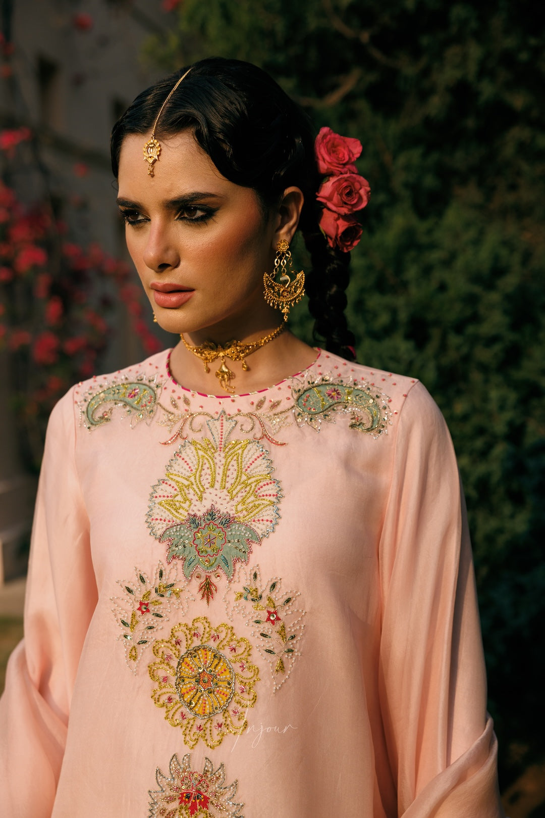A woman wearing a light pink embroidered traditional outfit with ornate gold jewelry and red roses in her hair, photographed outdoors against a dark green foliage background.