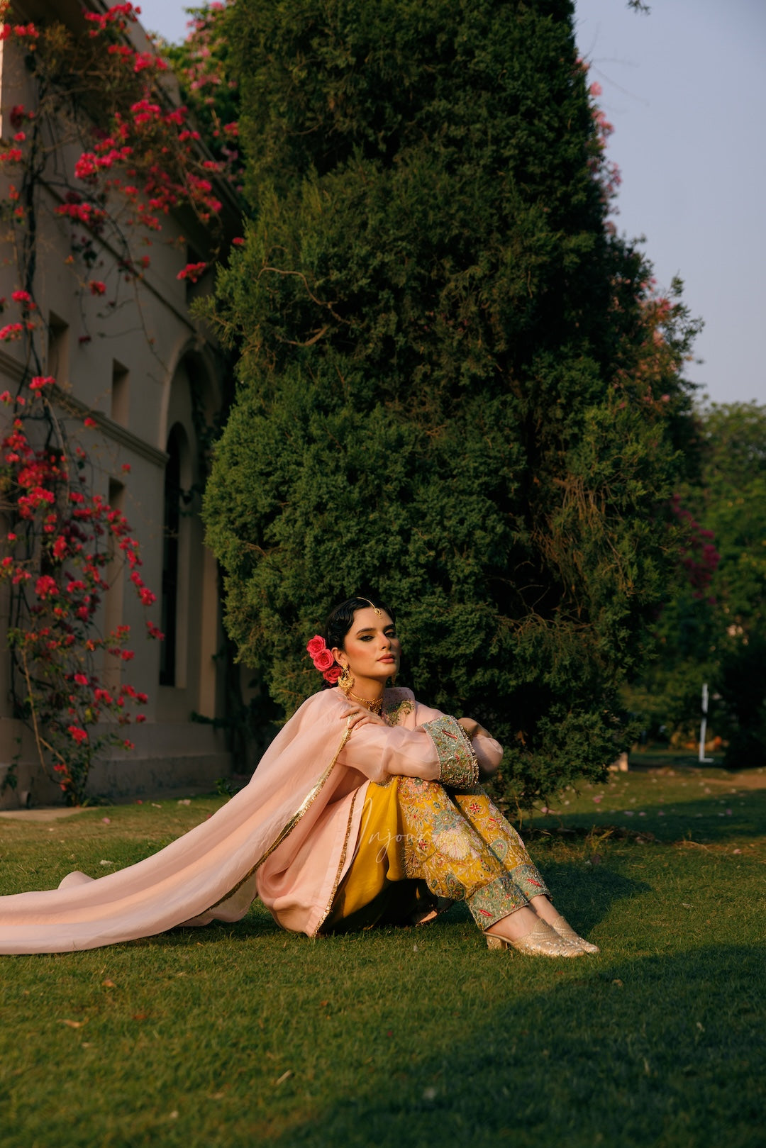 A woman in a traditional Indian outfit sits on the grass in front of a large green tree, wearing a light pink dupatta and a yellow patterned kurta with a pink floral hair accessory.