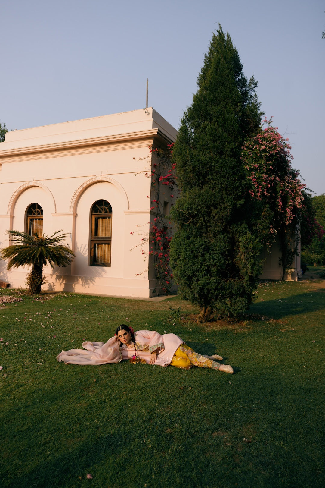 A woman in a light pink and yellow traditional outfit lies on a green lawn in front of a pale pink building with arched windows, a tall evergreen tree, and pink flowers.