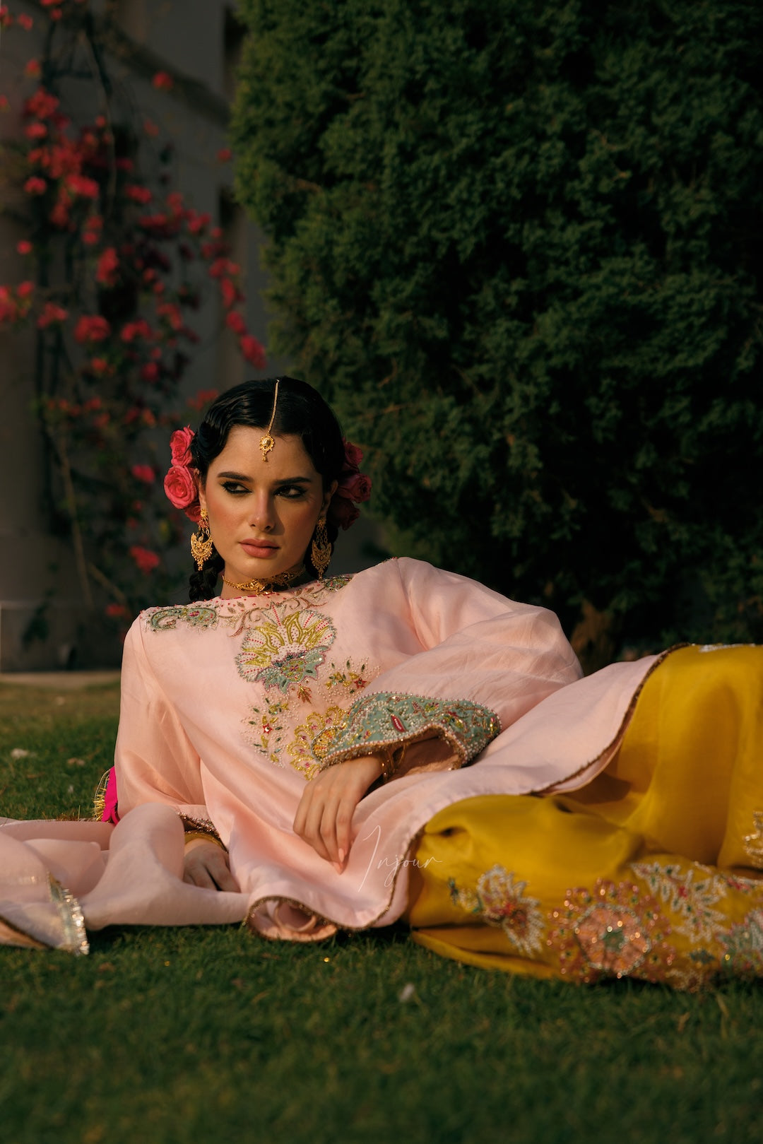 A woman reclines on grass outdoors wearing a light pink traditional outfit with colorful embroidered sleeves and a bright yellow skirt with matching embroidery. She has floral hair accessories and earrings, with a dark green hedge and flowering vines in the background.