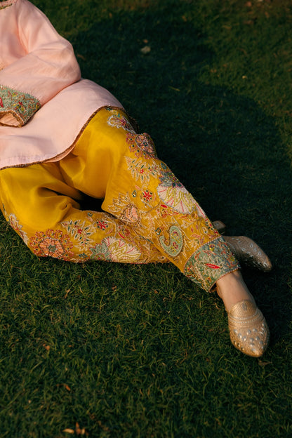 A person reclining on green grass, wearing a light pink traditional top and bright yellow embroidered pants with metallic embellishments and beige embellished shoes.