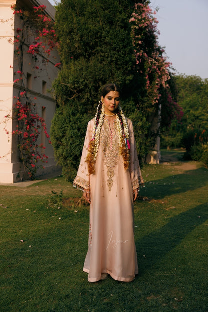 A woman stands outdoors on a grassy lawn wearing a long, light pink traditional gown with ornate gold embroidery and a matching embroidered headpiece. Trees, climbing pink flowers, and a building are visible in the background.