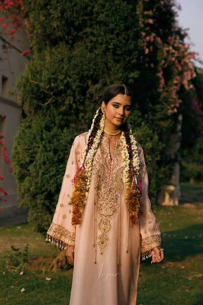 A woman wearing a traditional light pink embroidered kurta with gold detailing, floral garlands, and long braid hair stands outdoors in front of a large green tree.
