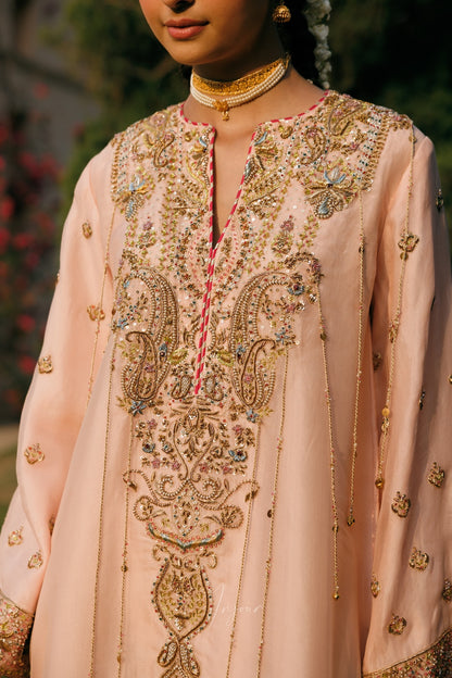 A close-up of a model wearing a light peach-pink traditional embroidered kurta with intricate gold, silver, and multicolor beadwork on the neckline and chest, paired with a gold choker necklace.