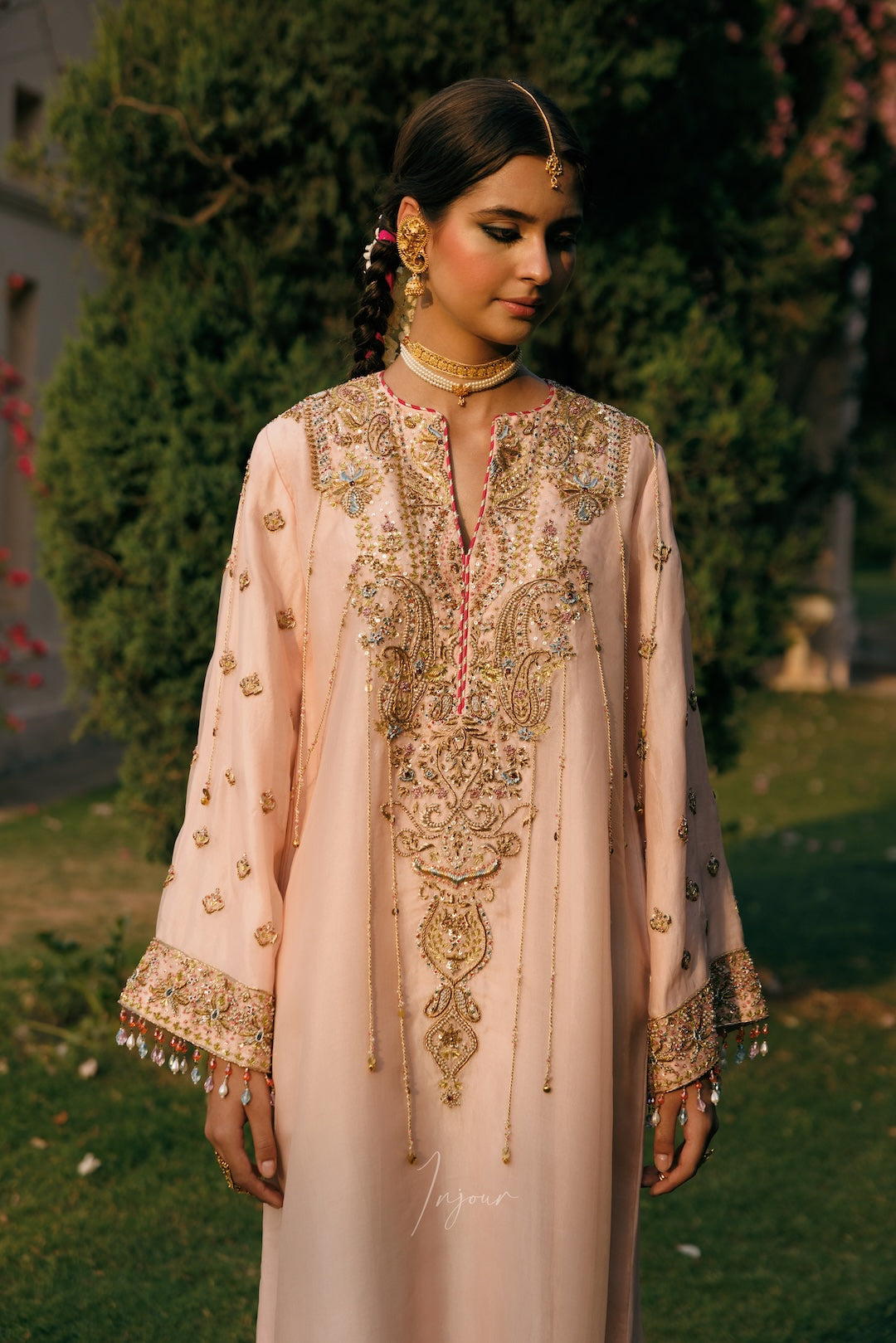 A woman wearing a light peach-pink traditional embroidered kurta with gold and multicolor beadwork, standing outdoors in front of green trees.
