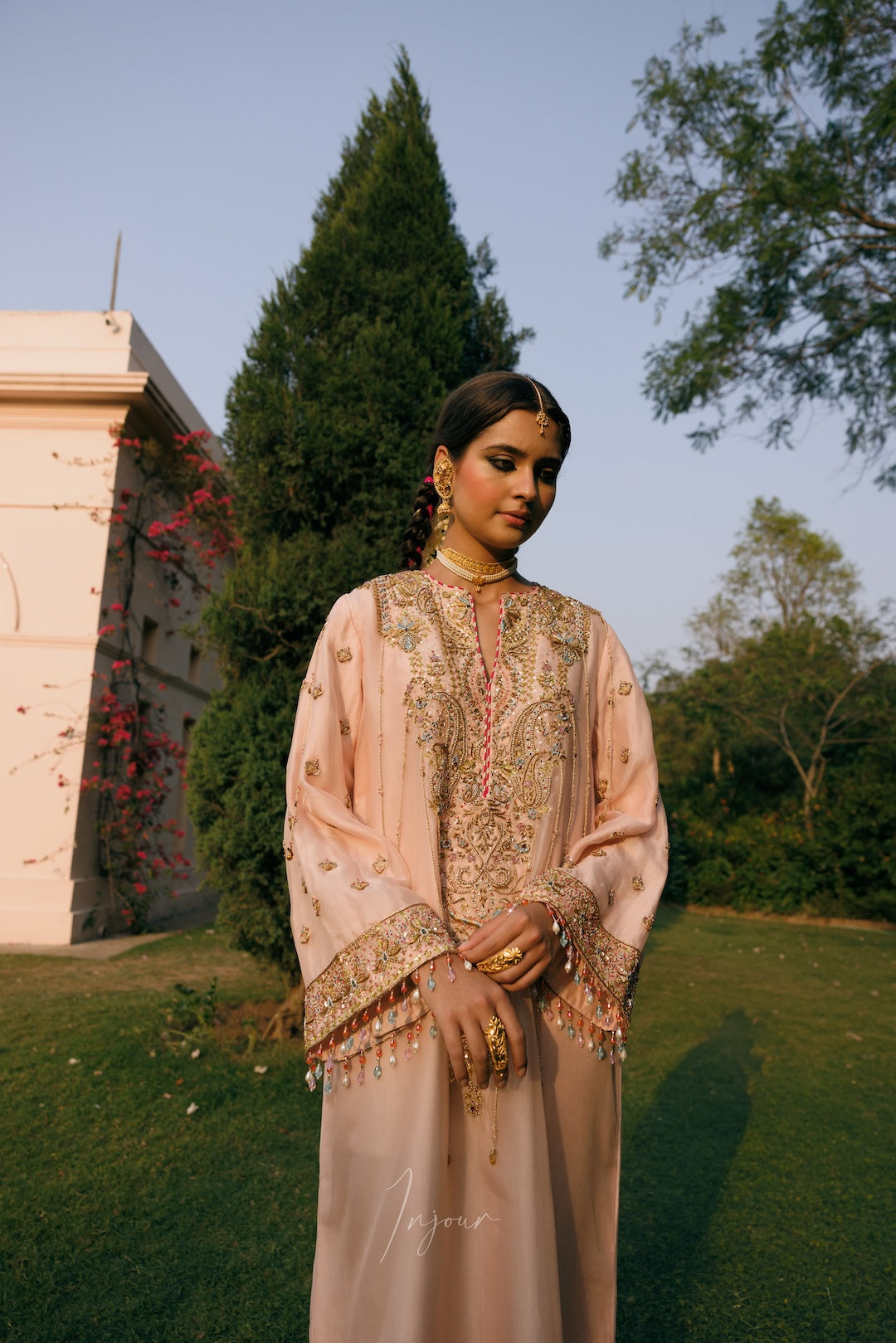 A woman stands outdoors in a garden wearing a light peach-beige traditional embroidered outfit with ornate gold detailing, layered jewelry, and a headpiece. Trees, grass, and a building are visible in the background.