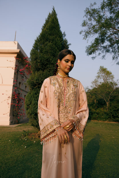A woman stands outdoors in a garden wearing a light peach-beige traditional embroidered outfit with ornate gold detailing, layered jewelry, and a headpiece. Trees, grass, and a building are visible in the background.