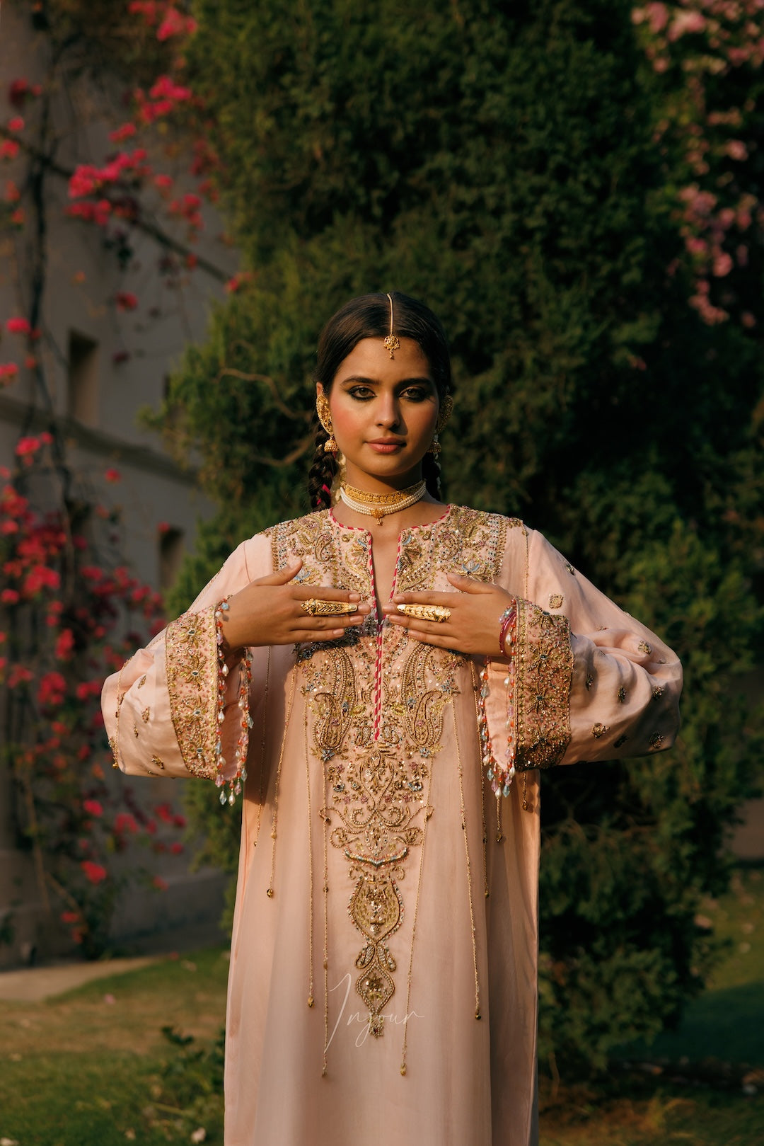 A woman wearing a light pink traditional embroidered kurta stands outdoors in front of a garden background with green foliage and pink flowers. The kurta features intricate gold embroidery and beadwork, and she is accessorized with gold jewelry.