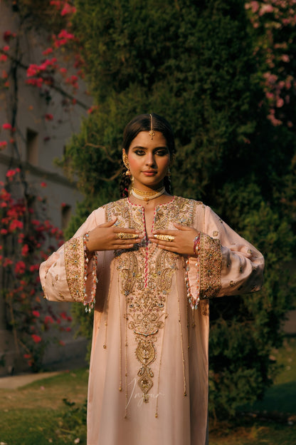 A woman wearing a light pink traditional embroidered kurta stands outdoors in front of a garden background with green foliage and pink flowers. The kurta features intricate gold embroidery and beadwork, and she is accessorized with gold jewelry.