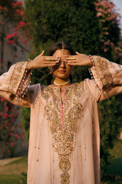 A woman wearing a light pink embroidered traditional outfit with ornate gold detailing, posing outdoors with her hands in front of her eyes against a background of green foliage and pink flowers.