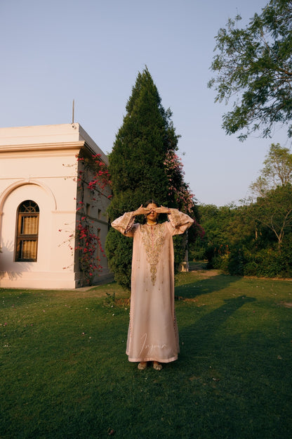 A woman wearing a long, light beige embroidered tunic stands on a grassy lawn in front of a white building and trees, with her hands on her head in a relaxed pose.